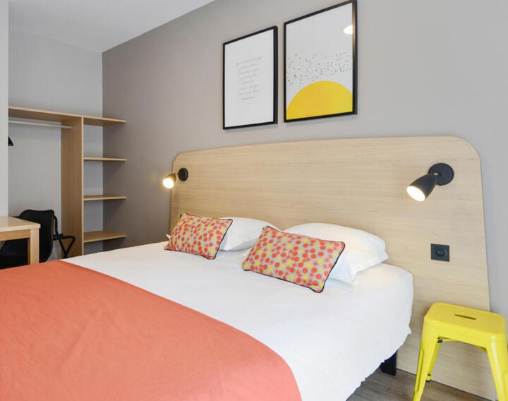 Modernes Zimmer in der AC Classic-Serie bei Appart'City mit einem Doppelbett mit gemusterten Kissen, einer orangefarbenen Decke, wandmontierten Leselampen, einem leuchtend gelben Stuhl, Holzregalen und abstrakter Wanddekoration, was einen stilvollen und funktionalen Raum schafft.