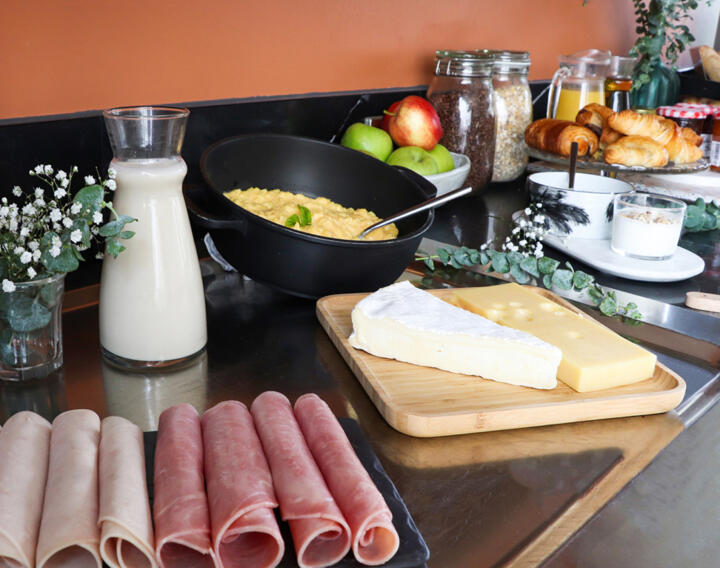 Breakfast buffet in an AC Classic aparthotel featuring slices of ham, cheese on a wooden board, scrambled eggs, a jug of milk, fresh fruit, cereals, and a selection of pastries on a black countertop.