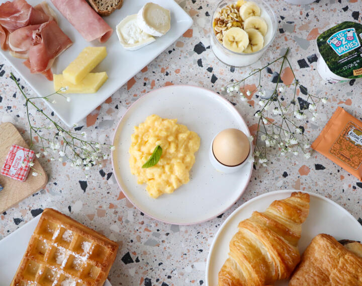 Petit-déjeuner gourmand disposé sur une table avec des œufs brouillés, un œuf à la coque, des viennoiseries, une gaufre, du fromage, du jambon, un bol de yaourt aux bananes, et des emballages de beurre et de confiture, pour un début de journée savoureux dans un appartement AC Confort.