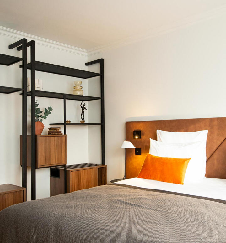 Habitación elegante de la gama Collection en Appart'City con una cama grande con cabecero de cuero marrón, cojines blancos y un almohadón naranja, estantes negros con objetos decorativos y un ambiente cálido y acogedor.