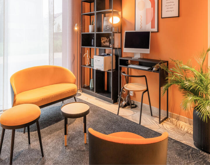 Cozy workspace in an Collection apartment with bright orange walls, a matching colored sofa and armchair, a round coffee table, a desk equipped with a computer, a bookshelf, and a green plant, creating a warm and inviting atmosphere.
