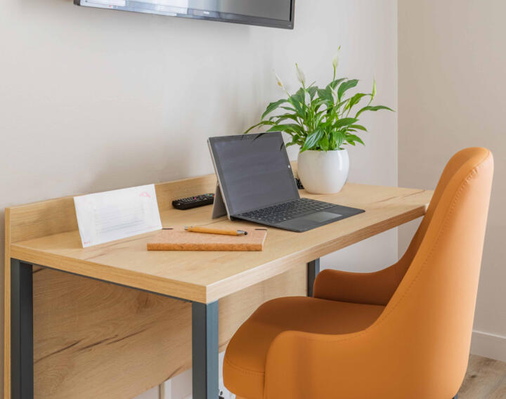 Organized workspace in an AC Confort apartment featuring a wooden desk, an ergonomic orange office chair, an open laptop, documents, and a potted green plant, providing a conducive setting for productivity in a comfortable environment.