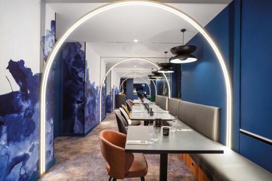 Vue du couloir du restaurant Bistrot City Genève Ferney-Voltaire avec des banquettes grises et des sièges en velours marron, sous un éclairage d'arches lumineuses, des murs bleus avec des taches abstraites et des luminaires suspendus noirs